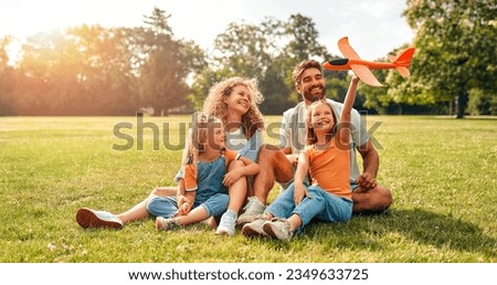Happy young family dad, mom and two children daughters playing with an airplane sitting in a meadow on the grass in the park on a warm sunny day, having fun on a day off.