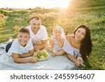 Happy young family eats fruits on vacation together. Father, mother, little girl, boy having fun on blanket in green grass, nature. Portrait of mom, dad, son, daughter. Concept of holiday on picnic.