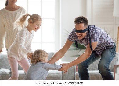 Happy young family of four playing hide and seek game at home, blindfolded father trying to catch children escaping, parents and kids having fun laughing spending free time together in living room