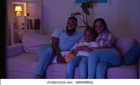 Happy Young Family Gathering Together In Front Of Tv Watching Movie In Evening. Positive Afro-american Mother, Father And Daughter Sitting Down On Sofa And Watching Tv In Dark Living Room