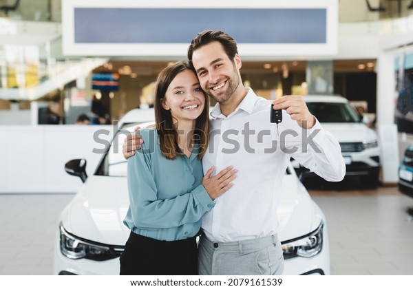 Happy young family holding car keys after buying new\
automobile at dealer shop store. Heterosexual husband and wife\
purchasing expensive auto