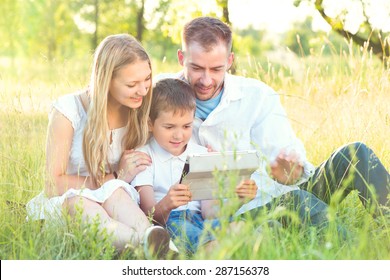 Happy Young Family with kid using Tablet PC in summer park. Dad, Mom and little boy with ipad computer resting outdoors together. Summer holidays
