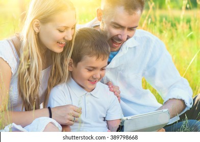 Happy Young Family With Kid Using Tablet PC In Summer Park. Dad, Mom And Little Boy With Computer Resting Outdoors Together. Summer Holidays