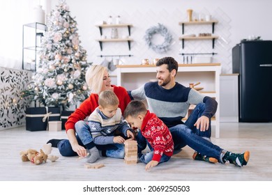 Happy Young Family With Kids In Woolen Jumpers Spending Winter Holidays At Home Playing With Wooden Blocks On Warm Floor In Cozy, Festively Decorated Living Room With Christmas Tree