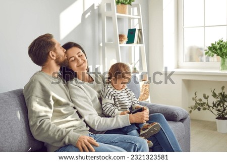 Happy young family with little child girl with smartphone sitting on the sofa. Husband kissing his wife with tenderness while their daughter watching funny cartoon online on mobile phone.
