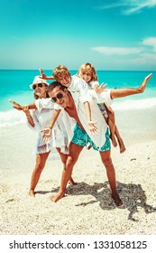 Happy Young Family With Little Kids Having Fun At The Beach. Mom And Dad Piggyback Their Sons.