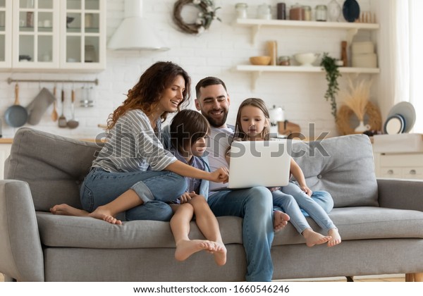 Happy young family with little kids sit on sofa in\
kitchen have fun using modern laptop together, smiling parents rest\
on couch enjoy weekend with small children laugh watch video on\
computer at home