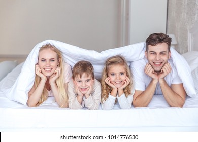 Happy Young Family Lying In Bed Together Under Blanket And Looking At Camera