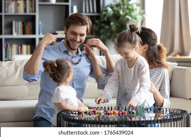 Happy young family making colorful wooden beads jewelry together, smiling father holding handmade necklace, excited mum and dad playing with two little daughters in living room, creative activity