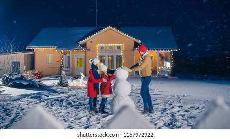 Happy Young Family Making Snowman in the Backyard of their Idyllic House. Father Puts Snowball, Daughter and Wife Help Him. Family Spending Time Together one Winter Evening.