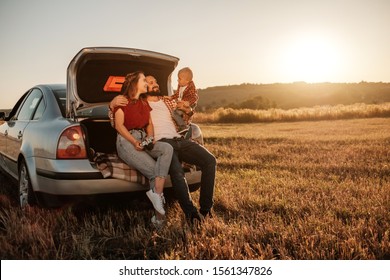 Happy Young Family Mom and Dad with Their Little Son Enjoying Summer Weekend Picnic Sitting on the Trunk of the Car Outside the City in the Field at Sunny Day Sunset, Vacation and Road Trip Concept