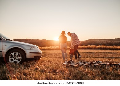 Happy Young Family Mom And Dad With Their Little Son Enjoying Summer Weekend Picnic On The Car Outside The City In The Field At Sunny Day Sunset, Vacation And Road Trip Concept