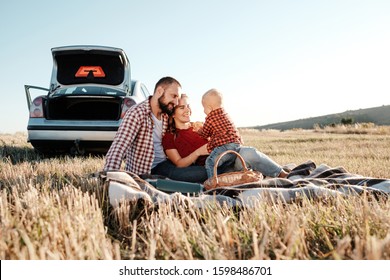 Happy Young Family Mom And Dad With Their Little Son Enjoying Summer Weekend Picnic Sitting On The Plaid Near The Car Outside The City In The Field At Sunny Day Sunset, Vacation And Road Trip Concept