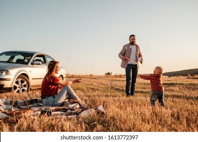 Happy Young Family Mom And Dad With Their Little Son Enjoying Summer Weekend Picnic On The Car Outside The City, Playing With Bubbles