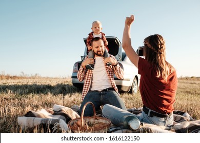 Happy Young Family Mom And Dad With Their Little Son Enjoying Summer Weekend Picnic Sitting On The Plaid Near The Car Outside The City In The Field At Sunny Day Sunset, Vacation And Road Trip Concept