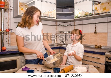 Happy young family, mother and daughter, having fun preparing cookies on light European kitchen, mom shows and teaches daughter how to correctly knead the dough and roll it out