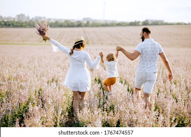 Happy Young Family Mother Father And Son Walk On Nature On Sunset Hold Hand. Back View. Parents Playing With Their Child Outside In A Field With Pink Sage Flower. Space For Copy. From Behind.