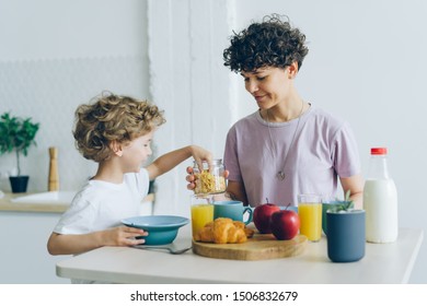 Happy Young Family Mother And Little Son Eating Cereal With Milk At Home Talking Enjoying Domestic Breakfast In Cosy Kitchen. People And Meals Concept.