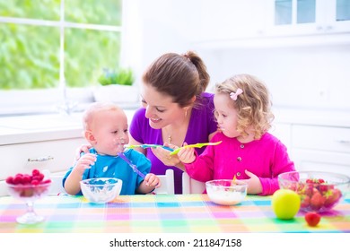 Happy young family, mother with two children, adorable toddler girl and funny baby boy having healthy breakfast eating fruit and dairy, sitting in a white sunny kitchen with window