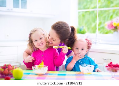 Happy Young Family, Mother With Two Children, Adorable Toddler Girl And Funny Messy Baby Boy Having Healthy Breakfast Eating Fruit And Dairy, Sitting In A White Sunny Kitchen With Window