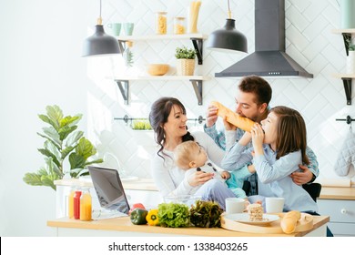 Happy Young Family With Mum, Dad And Two Different Age Children Cooking Breakfast, Laughing Biting The Baguette In The Kitchen Preparing Meal Together.