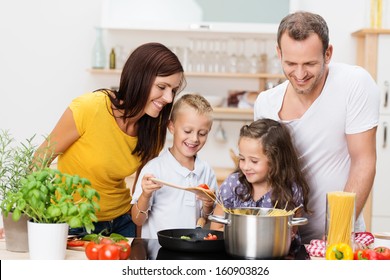 Happy Young Family With Mum, Dad And Two Young Children Cooking In The Kitchen Preparing A Spaghetti Meal Together