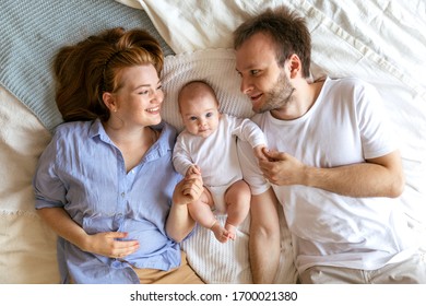 Happy Young Family With A Newborn Baby Lying On The Bed, Smiling And Looking At The Camera. How Life Changes With The Advent Of A Child