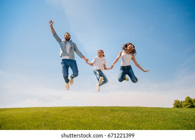 Happy Young Family With One Child Holding Hands And Jumping On Meadow