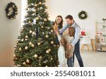 Happy young family parents and children gathering together on Christmas, beautifying their house. Joyful father, mother and two kids preparing to place golden star on top of decorated Christmas tree.