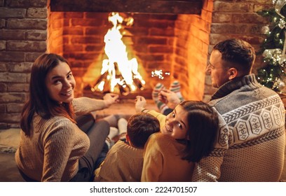 Happy young family parents with two small kids gathering around brick fireplace in cozy and warm country house during Christmas holidays in winter. Father, mother ad children sitting by fire at home - Powered by Shutterstock