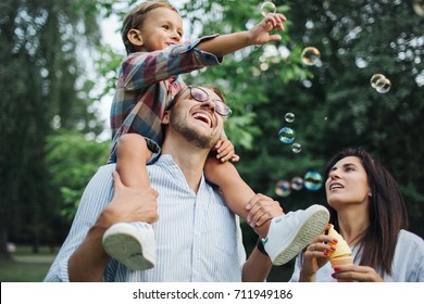 Fröhliche junge Familie, die mit Blaswandeln spielt, mit Sohn, der im Sommer im Park im Freien auf den Schultern der Väter sitzt