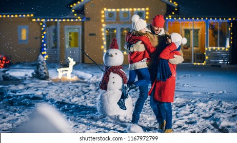 Happy Young Family Portrait In The Falling Snow, Father Embraces Wife And Holds Daughter. Family Enjoying Winter Holiday In The Front Yard Of Their Idyllic House Decorated With Garlands.