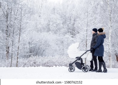 Happy young family pushing white baby stroller and walking at nature park. Snow covered trees and bushes. Spending time with infant in beautiful winter day. Enjoying peaceful stroll. Side view. 