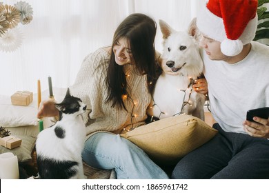 Happy young family sitting with cute dog and cat in festive decorated boho room, sweet moments. Merry Christmas! Stylish couple with pets in lights and santa hat celebrating holidays at home