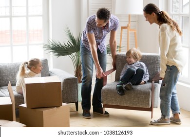 Happy young family with small kids having fun together playing on moving day in new home concept, father and mother carry little son on chair while excited daughter sitting on sofa unpacking boxes