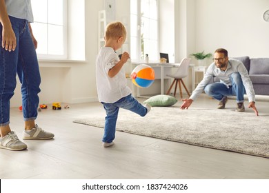 Happy Young Family Staying At Home, Having Fun Together And Engaging Their Small Kid In Sport Games. Dad With Little Son Playing Soccer With Inflatable Ball In Cozy Living-room