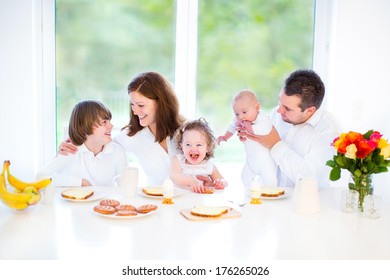 Happy Young Family With A Teenage Boy, Adorable Curly Toddler Girl And A Newborn Baby Having Fun Together On A Sunday Morning Having Breakfast In A White Dining Room With A Big Window 