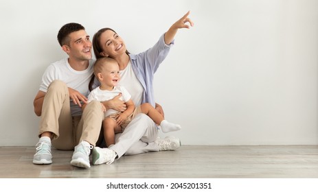 Happy Young Family Of Three With Infant Baby Sitting On Floor Near White Wall And Pointing Aside At Copy Space, Parents And Child Demonstrating Free Place For Your Design Or Advertisement, Panorama