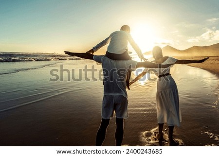 Happy young family of three spending free time outdoors, walking and having fun on seaside. Back view. Copy space. Island vibes. Sunset light. Vacation.
