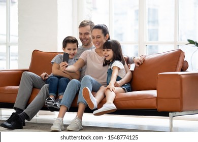 Happy Young Family With Two Kids Sit On Couch Making Self-portrait Picture On Smartphone Together, Smiling Parents And Children Have Video Call Use Fast Unlimited Wireless Internet Connection On Cell