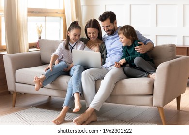 Happy young family with two small kids relax on sofa in living room at home have fun using laptop together. Smiling Caucasian parents with little children talk speak on webcam video call on computer.