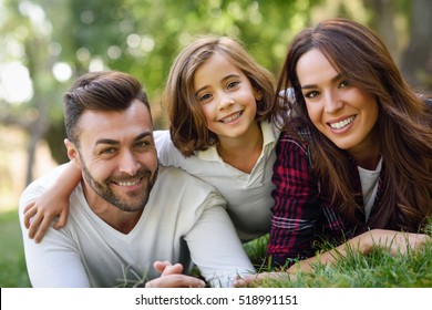 Happy Young Family In A Urban Park. Father, Mother And Little Daughter Laying On Grass.