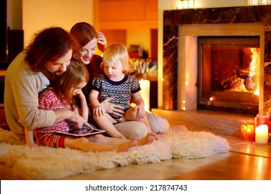 Happy young family using a tablet pc at home by a fireplace in warm and cozy living room on winter day