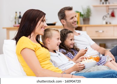 Happy young family watching television with attractive young parents sitting with their two children on a sofa in the living room laughing at the program