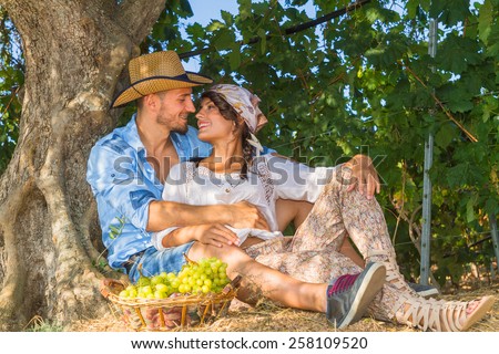 Happy young farmers enjoy a work break sitting at the shade of a tree at the vineyard, eating grapes, kissing, feeding, and teasing one another.