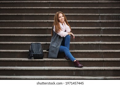 Happy young fashion woman with handbag sitting on steps Stylish female model in gray sleeveless coat and light pink sweater