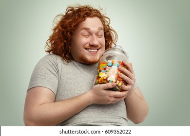 Happy young fat obese man smiling joyfully, keeping eyes closed rejoicing at glass jar of goodies, can't wait to start eating unhealthy but so tasty sweets while keeping strict vegetable diet