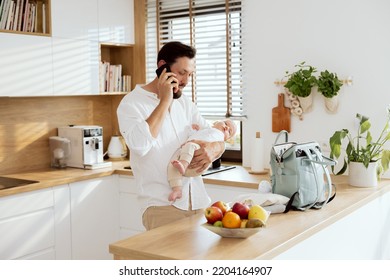 Happy Young Father Dad In Moderln Light Kitchen Holding Adorable Baby In Arms Talking By Phone Mobilephone With Wife. On Wooden Surface Backpack With Milk Bottle Baby Meal.