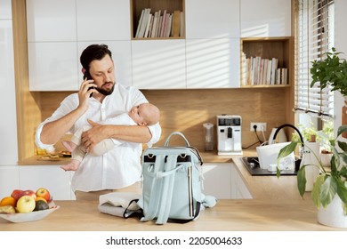 Happy Young Father Dad In Moderln Light Kitchen Holding Adorable Baby In Arms Talking By Phone Mobilephone With Wife. On Wooden Surface Backpack With Milk Bottle Baby Meal.