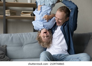 Happy Young Father Lifting In Air Upside Down Laughing Adorable Small Kid Son, Having Fun Together In Living Room. Joyful Two Male Generations Family Enjoying Entertaining Activity On Weekend At Home.